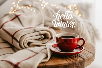Cozy winter scene with a red cup of coffee and a warm blanket on a wooden table with soft bokeh lights creating a relaxing and inviting atmosphere.