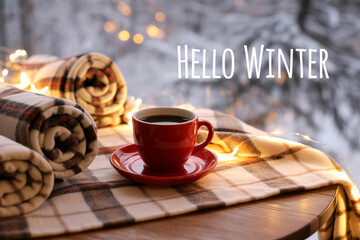 Cozy winter scene with a red cup of coffee and warm blankets on a wooden table against a snowy backdrop creating a feeling of comfort and relaxation.