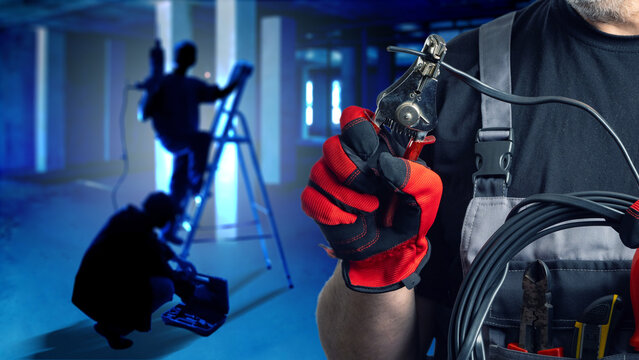 Electrical installation team performs wiring work, with a technician holding cable cutters while colleagues set up lighting and equipment in a building.