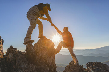 Asia couple hiking help each other.Silhouette Two Male hikers climbing up mountain cliff and one of them giving helping hand. People helping and, team work concept.