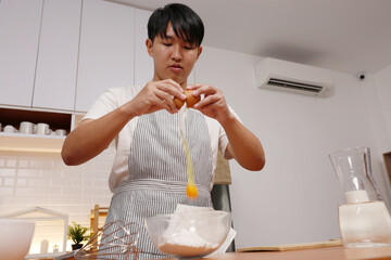 Asian Young person in striped apron cracking egg into mixing bowl while preparing batter in modern kitchen, focused and calm
