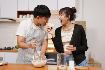 Asian young couple baking in kitchen feeding each other sandwich and laughing while whisking batter and holding milk