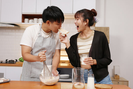Asian young couple baking in kitchen feeding each other sandwich and laughing while whisking batter and holding milk