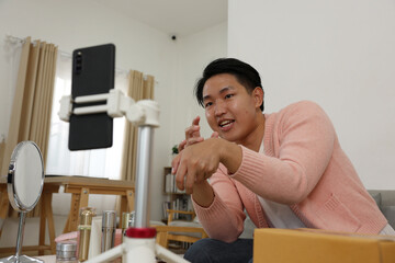 Asian young man recording beauty tutorial with skincare products on table smiling and demonstrating application
