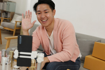 Asian Young man waving and smiling during live video stream with skincare bottles and shipping boxes on table