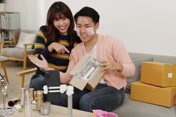 Asian Young couple unboxing skincare set with sheet masks smiling during live stream, beauty products and cardboard boxes on table, cozy living room scene