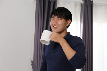 Asian young man smiling with eyes closed holding coffee mug in living room, casual morning moment, relaxed expression, indoor portrait
