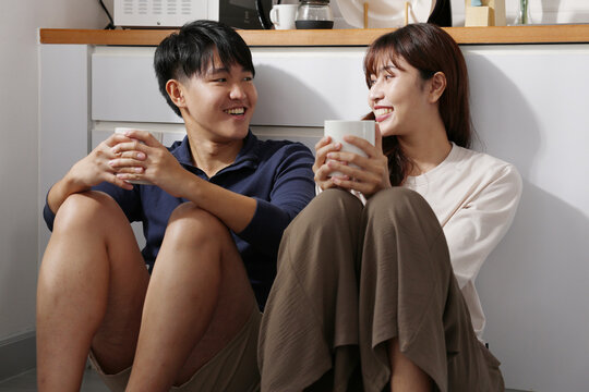 Asian Young couple sitting on kitchen floor holding mugs smiling and relaxed while sharing warm conversation and casual morning drink together - Powered by Adobe