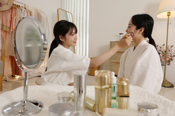 Asian two women in bathrobe applying makeup in bedroom mirror, skincare routine, smiling friends sharing beauty tips, cozy dressing area with lamp and clothes rack