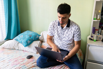 Young man working remotely with laptop while sitting on his bed in a cozy and personal bedroom space