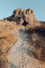 Hiking trail winding through high plains scrub including sagebrush and dappled by late afternoon shade, to large rock formation under a clear blue sky. 
