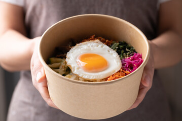 Bibimbap, Korean food, Cooked rice with vegetables, pork and egg in biodegrable bowl holding by hand