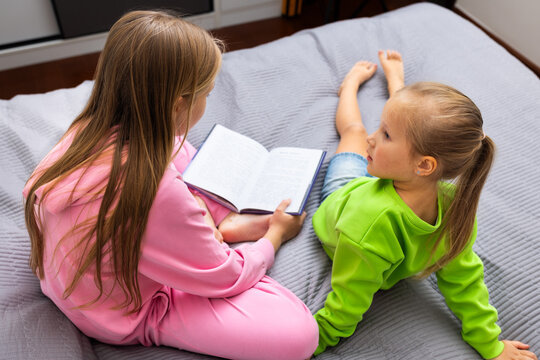 Teen girl reading a book to a young girl while sitting on a bed at home