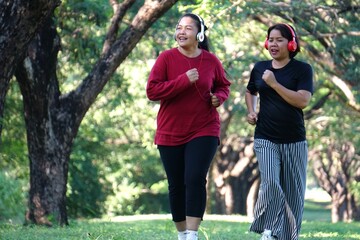 Two women are jogging side by side along a shaded path in the park, both smiling and looking forward.