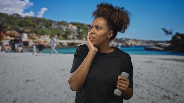 Young african american woman grimacing with hand on cheek while holding a small dumbbell on a crowded sandy beach by the sea; pain toothache.
