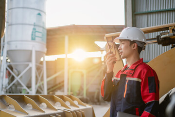 An engineer standing at industrial material storage yard checking data plans on laptop. Project management quality control. Large cement silo and pile of materials in background.