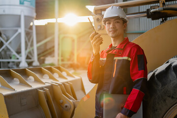 A young construction engineer leaning against the scoop of a large bulldozer in an industrial area, operating heavy equipment, project within a material processing or construction environment.