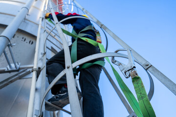 A worker or engineer, wearing essential safety equipment including a harness and a yellow hard hat, is captured climbing a fixed vertical ladder on the side of a large industrial silo.