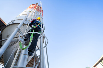 A worker or engineer, wearing essential safety equipment including a harness and a yellow hard hat, is captured climbing a fixed vertical ladder on the side of a large industrial silo.