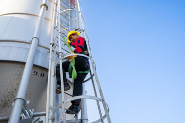 A worker or engineer, wearing essential safety equipment including a harness and a yellow hard hat, is captured climbing a fixed vertical ladder on the side of a large industrial silo.