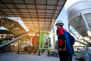Two construction or factory engineers, wearing protective gear, stand inside an industrial processing plant. One is focused on a laptop, while the other communicates via a walkie-talkie.