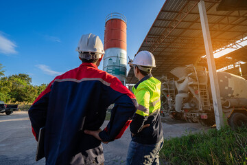 Two construction engineers wearing hard hats stand looking at a large cement silo in an industrial yard, pointing at the structure and discussing plans with colleagues.