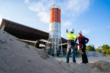 Two construction engineers wearing hard hats stand looking at a large cement silo in an industrial yard, pointing at the structure and discussing plans with colleagues.