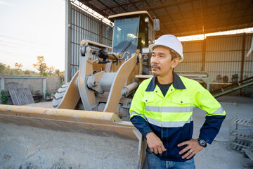 A Professional Asian site manager stands confidently with hands on hips in front of a large yellow wheel loader or heavy machinery in a material handling shed.