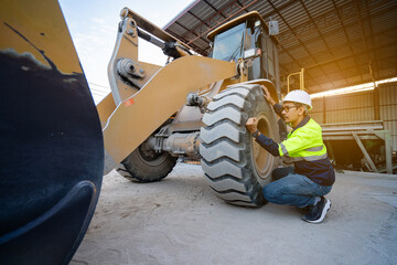 Asian male engineer wearing protective gear climbs into the passenger compartment of a large yellow wheel loader. Heavy machinery and workers working inside a material storage shed.