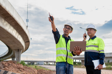 Two construction engineers, wearing hard hats and high-visibility vests, stand at a highway interchange construction site. discussing plans with his colleague who holds a walkie-talkie and tablet.