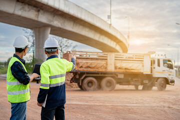 Two construction engineers, wearing hard hats and high-visibility vests, stand at a highway interchange construction site. discussing plans with his colleague who holds a walkie-talkie and tablet.