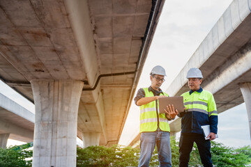 Two construction engineers, wearing hard hats and high-visibility vests, stand at a highway interchange construction site. discussing plans with his colleague who holds a walkie-talkie and tablet.