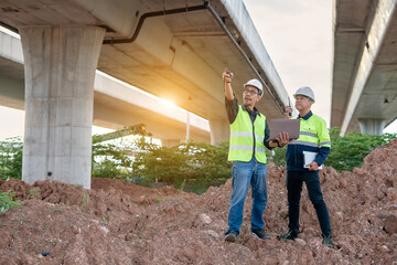 Two construction engineers, wearing hard hats and high-visibility vests, stand at a highway interchange construction site. discussing plans with his colleague who holds a walkie-talkie and tablet.