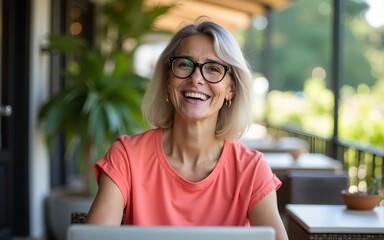 Portrait of confident mature professional woman in glasses, a coral T-shirt sitting on summer terrace in cafe, using laptop computer for work, laughing happily indoors. High quality