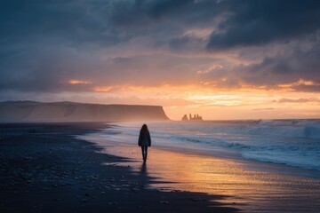 Fototapeta premium Woman strolls along a black sand beach in Iceland during a vibrant sunset with waves gently crashing in the background