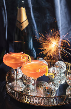 Two orange cocktails on a gold tray with small disco balls, champagne, and a lit sparkler for a New Year's Eve celebration. 