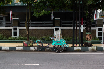 Blue traditional pedicab parked on a sidewalk near a street with trees. Indonesian traditional transportation vehicle.