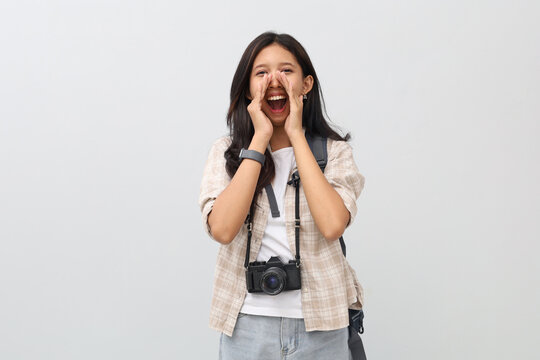 A young Asian woman wearing casual clothes, backpack, camera hanging around her neck, shouting sharing news or announcement with her hand near her mouth, vacation concept.