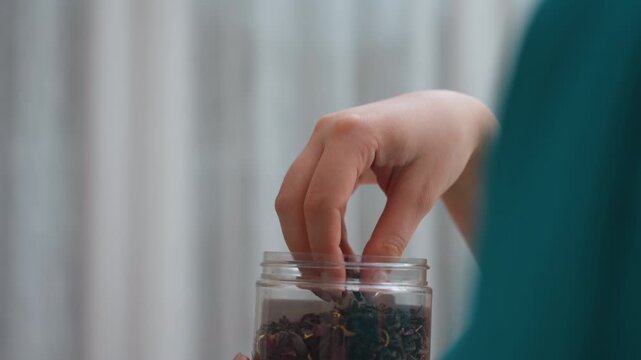 Caucasian Barista Selecting Tea Leaves From Jar Poised Hand Dips Into Glass Container To Choose Leaves, Teal Uniform Visible, Refined Careful Motion For Brewing, CloseUp Captures Texture And Aroma