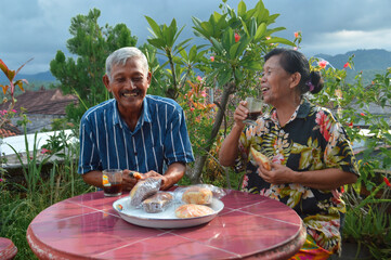 A joyful shot of an elderly couple at a red outdoor table surrounded by flowers, laughing together while enjoying coffee in glass mugs and pastries in the afternoon sunshine.