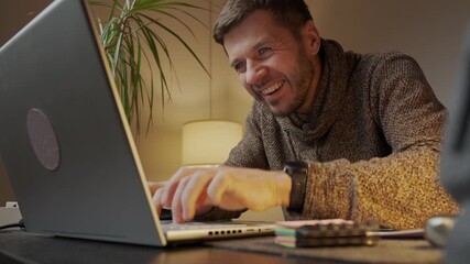Maliciously smiling man sitting at home office desk rubbing his hands and typing on laptop keyboard. Person writes comments and provokes others on internet. Internet troll and online behavior concept