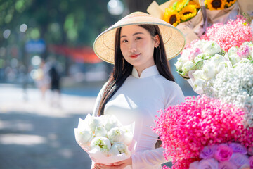 A young Vietnamese woman wearing a white ao dai and a conical hat stands beside a colorful street...