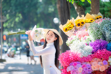 A young Vietnamese woman wearing a white ao dai and a conical hat stands beside a colorful street...