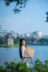 A young Vietnamese woman in a white ao dai and a conical hat standing by Hoan Kiem Lake in Hanoi, Vietnam