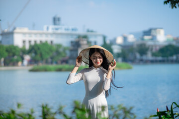 A young Vietnamese woman in a white ao dai and a conical hat standing by Hoan Kiem Lake in Hanoi, Vietnam