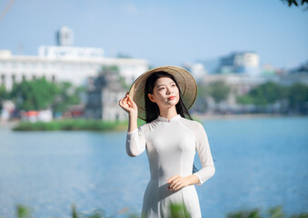 A young Vietnamese woman in a white ao dai and a conical hat standing by Hoan Kiem Lake in Hanoi, Vietnam