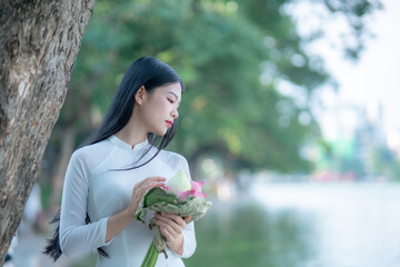 A young Vietnamese woman in a white ao dai standing by Hoan Kiem Lake, holding a bouquet of pink lotus flowers