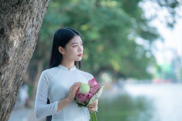 A young Vietnamese woman in a white ao dai standing by Hoan Kiem Lake, holding a bouquet of pink lotus flowers