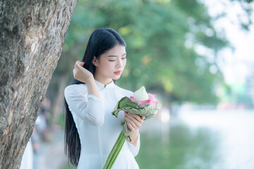 A young Vietnamese woman in a white ao dai standing by Hoan Kiem Lake, holding a bouquet of pink lotus flowers