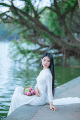 A young Vietnamese woman in a white ao dai standing by Hoan Kiem Lake, holding a bouquet of pink lotus flowers